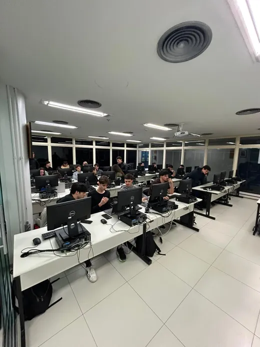 Fileiras de bancadas com estudantes programando em desktops durante encontro da Liga de Algoritmos